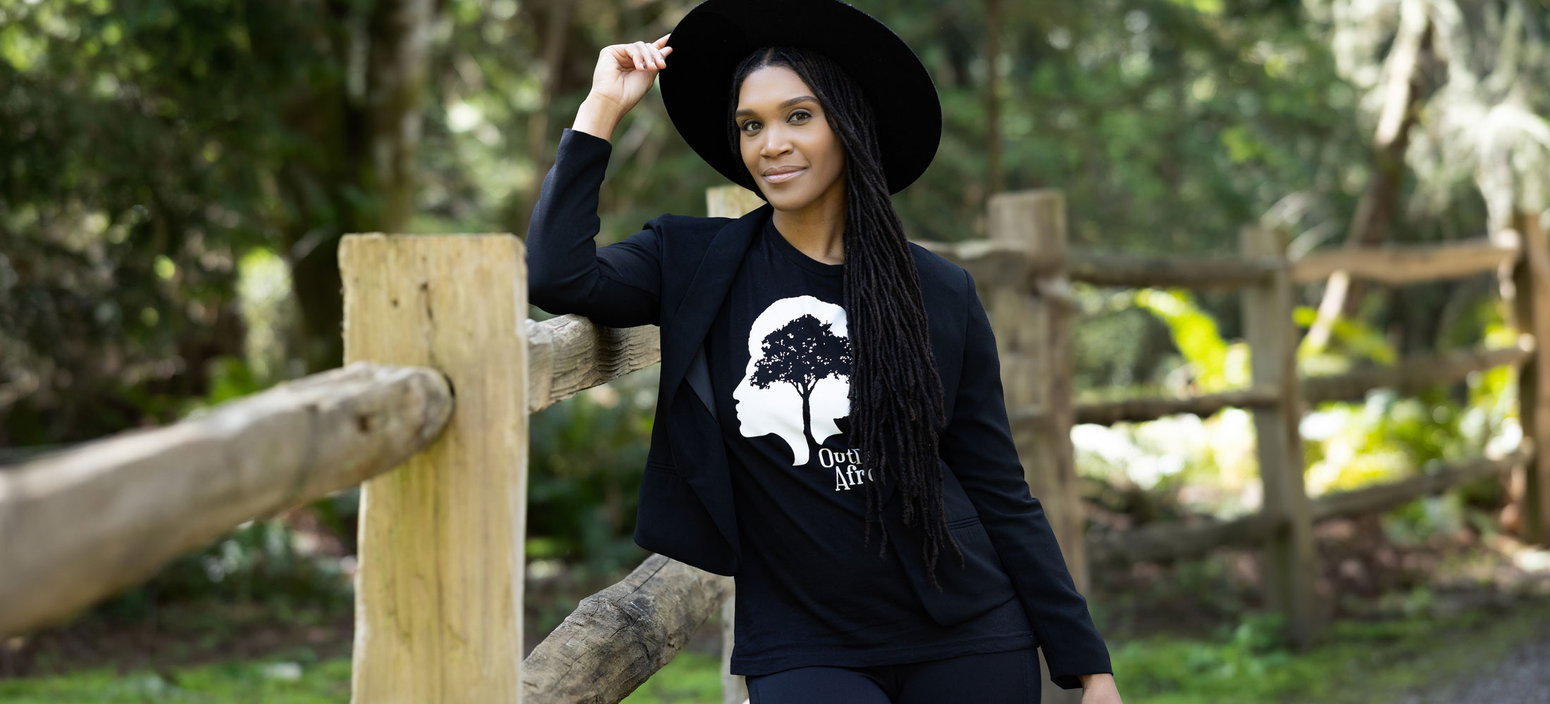 Woman wearing a black shirt and a black hat leaning against a fence with her hand on her hat, posing.